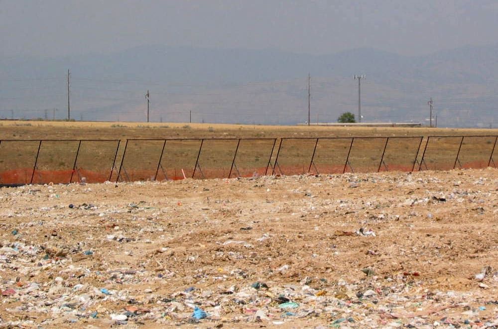 A landfill view showing a large, littered and barren field with scattered trash, a row of portable litter fences in the background, and power lines and mountains in the distance.