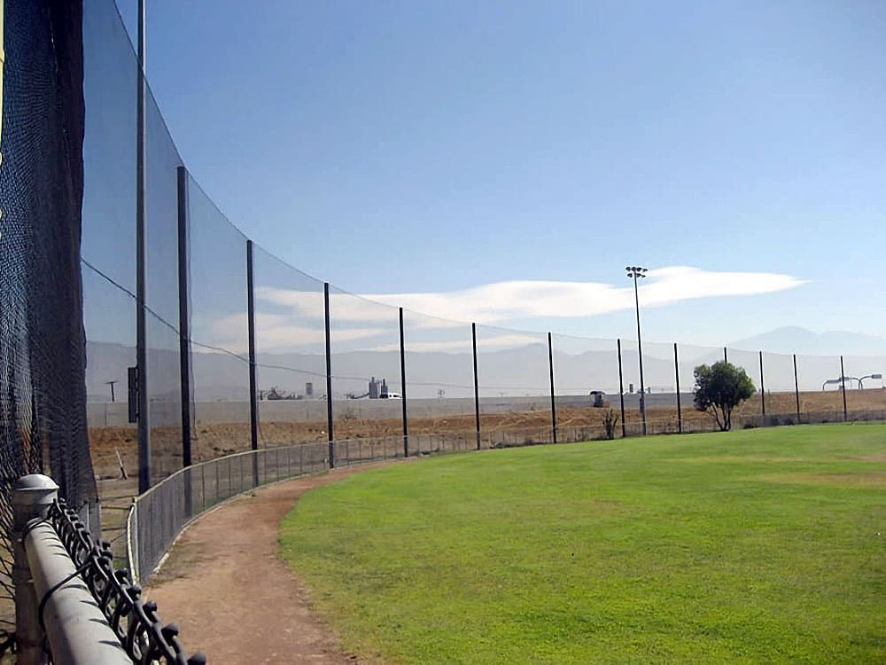 A baseball field with a green grass infield is bordered by a high netting system with steel poles. The background features a dry landscape and distant mountains under a clear blue sky.