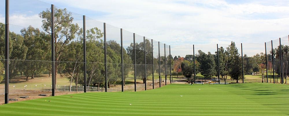 A grassy driving range with tall netting system and surrounding trees under a partly cloudy sky.