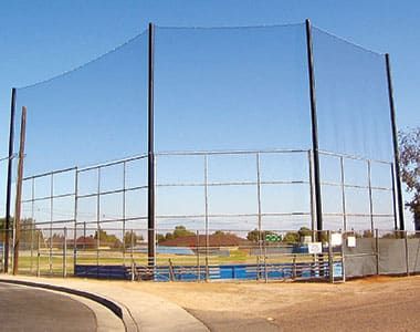 A baseball field with a backstop net and surrounding fence, positioned near a curved road. The field is empty and the sky is clear.