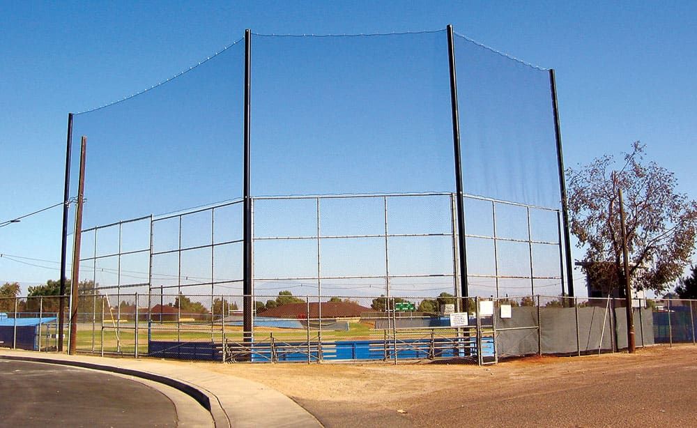 A baseball field with a backstop net and surrounding fence, positioned near a curved road. The field is empty and the sky is clear.