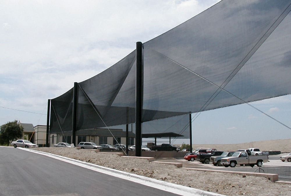 Netting structure extending over a parking area with multiple cars parked underneath. The framework is supported by tall black steel poles. The scene is set on a slightly inclined road on a clear day.