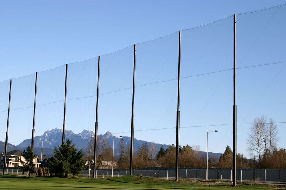 A tall golf driving range netting system with several supporting steel poles stands against a blue sky and distant mountains, with grass and trees in the foreground.