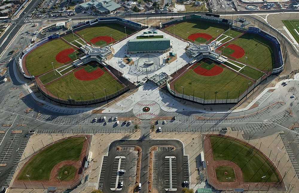 Aerial view of a sports complex featuring four baseball fields with red infields and a central building, surrounded by netting fences protecting adjacent parking lots and roads.