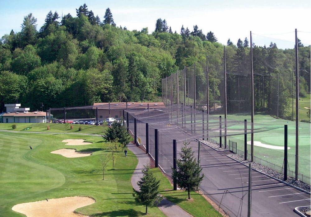 A road and parking lot enclosed by netting, protecting it from the golf course with sand bunkers on the left and a fenced driving range on the right, bordered by a forested area in the background.