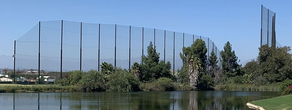 Tall netting surrounds a golf course fairway with trees near a water body under a clear blue sky.