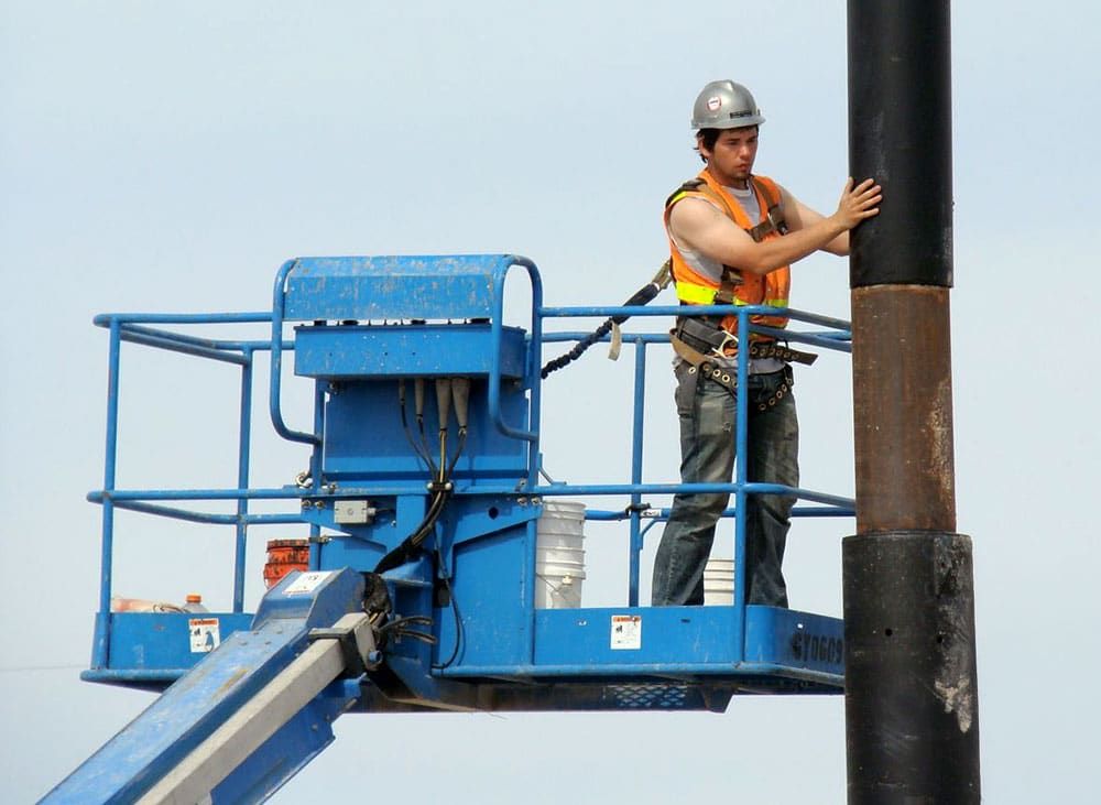 Construction worker in a safety vest operates a blue cherry picker while securing a large Coastal steel netting pole installation..