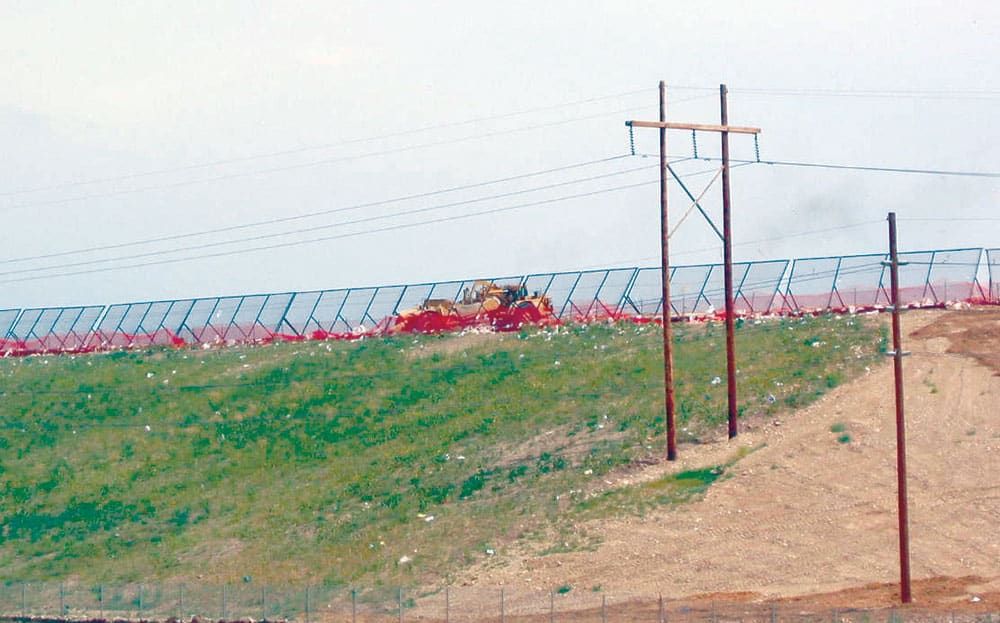 A bulldozer operates near a long portable litter fence on a grassy embankment, with telephone poles and electrical wires in the foreground.