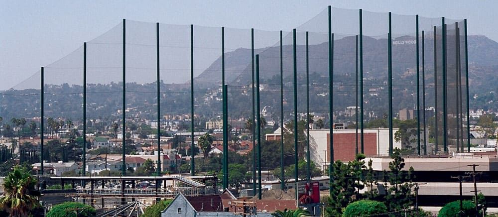 Tall safety nets surround a golf driving range, with the Hollywood Hills and its iconic sign visible in the background.