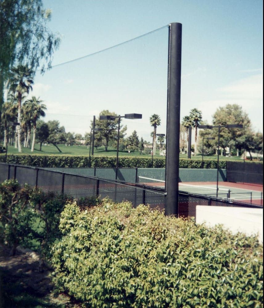 Outdoor tennis court surrounded by a netting fence and greenery, with trees, shrubs and grass and a clear sky in the background.