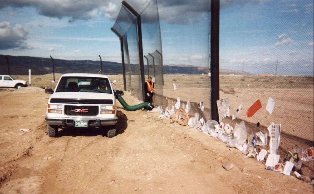 A white truck next to a person collecting trash that has collected along the base of a stationary litter fence in a desert landscape. Cloudy sky overhead.