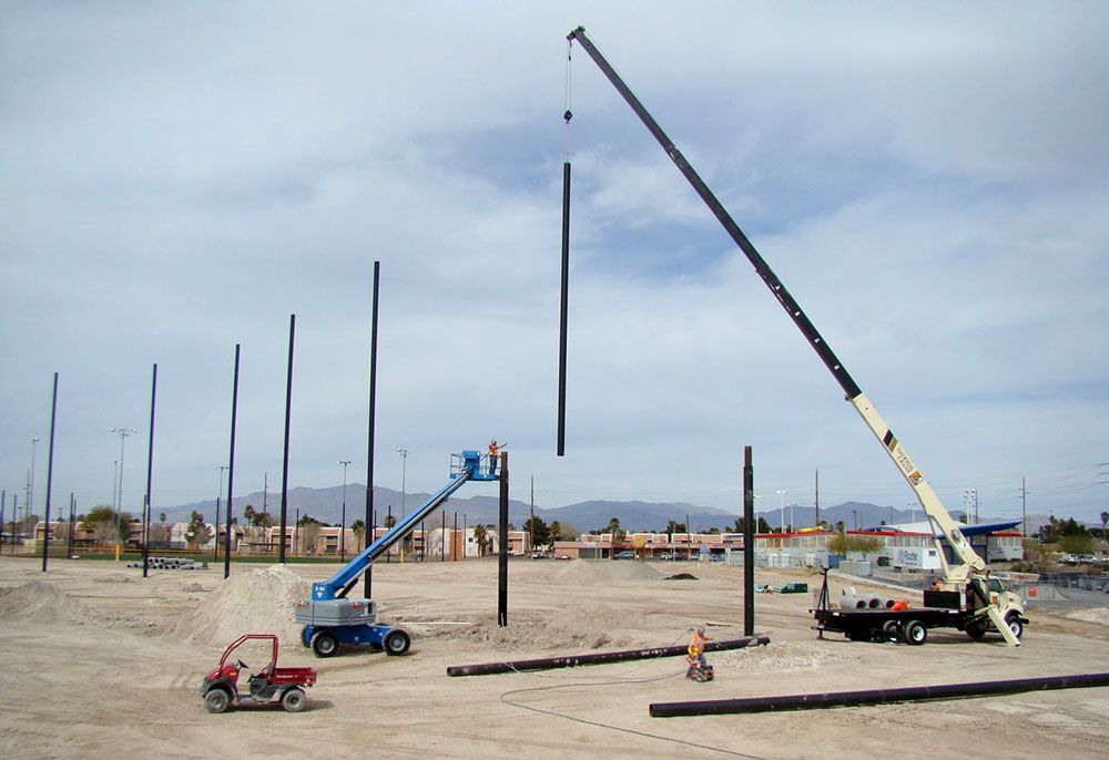 Construction site with cranes lifting and installing tall Coastal steel poles. Workers operate machinery on a flat, dirt terrain under a cloudy sky with distant buildings and mountains visible.