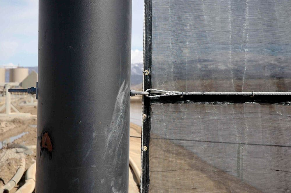 A close-up of a black steel pole supporting a dark netting fence. The background shows an industrial area with blurry structures and pipes.