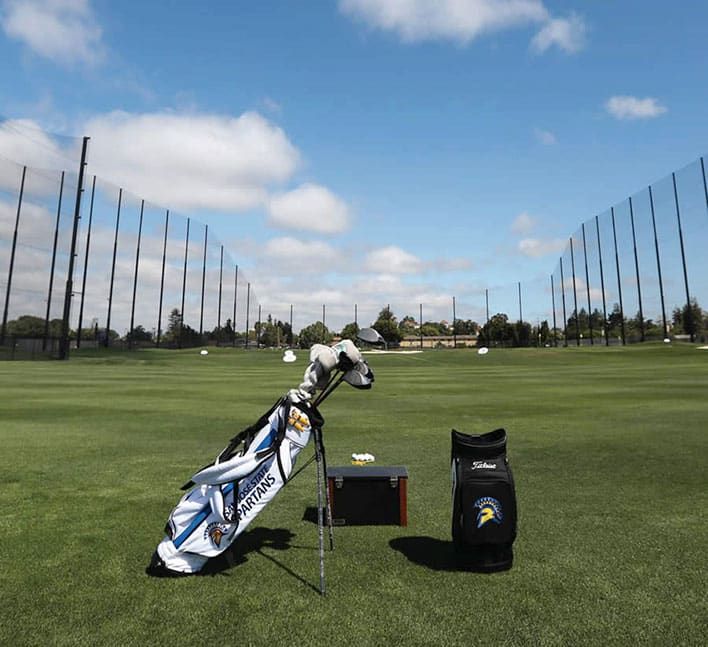 Golf course practice area with two golf bags, a small bench, and some scattered golf balls. The background includes a driving range netting system and trees under a partly cloudy sky.