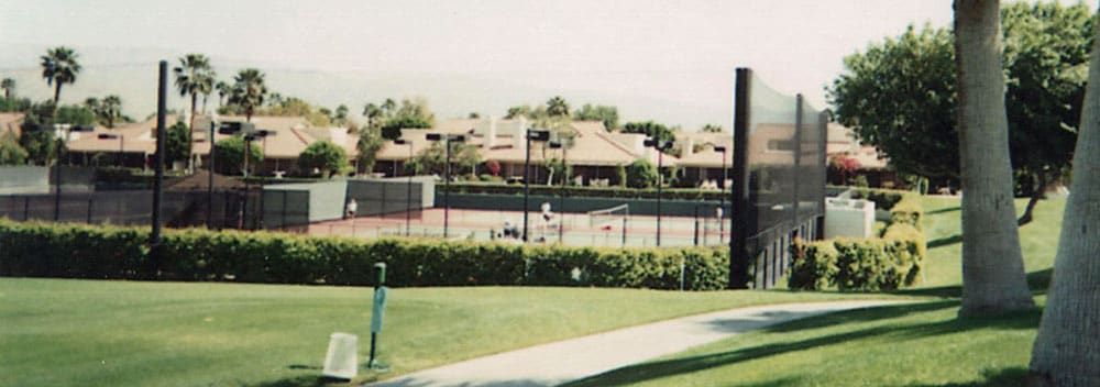 Tennis courts surrounded by protective netting system and hedges with players in action, set against a backdrop of houses and palm trees on a sunny day.