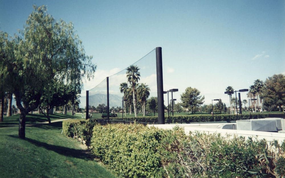 Black poles support a netted barrier beside a tennis court, screening the court from a green park with trees, hedges, lawn, and a cement path. Mountains and a clear blue sky are visible in the background.
