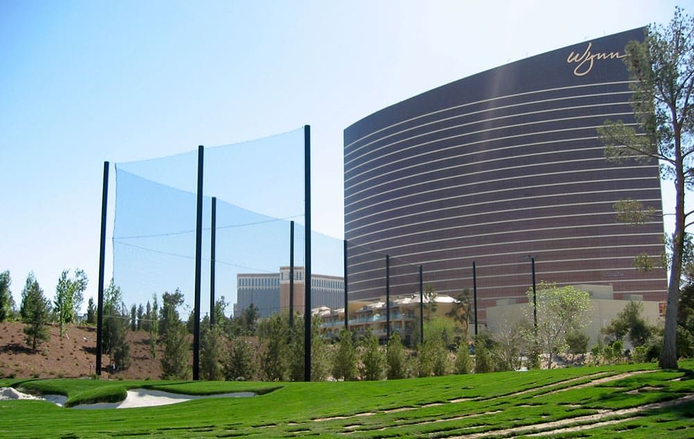 Golfers enjoy a lush green golf course by a serene pond, with an advanced golf course netting system standing tall in the background. Two golf carts rest nearby under the clear blue sky.