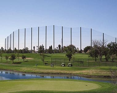 A well-maintained driving range with several tall steel poles and netting in the background. The sky is partly cloudy, and mountains are visible in the distance.