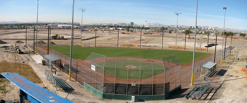 A baseball field surrounded by construction areas, with a cityscape visible in the background. The field includes green grass, bases, and a pitcher's mound, all enclosed by a netting systerm fence.