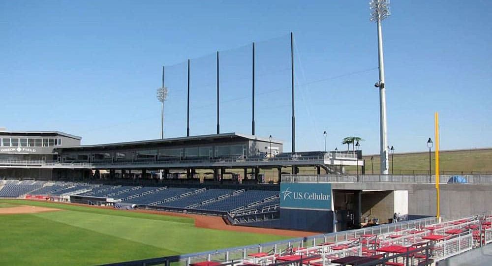 Empty baseball stadium with tall baseball netting system, rows of red seats, a green field, overhead lights, and a US Cellular advertisement on the structure in the background.