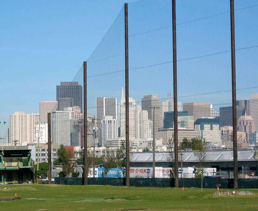 A driving range with tall netting is in the foreground, and a city's skyline featuring multiple high-rise buildings is in the background under a clear blue sky.