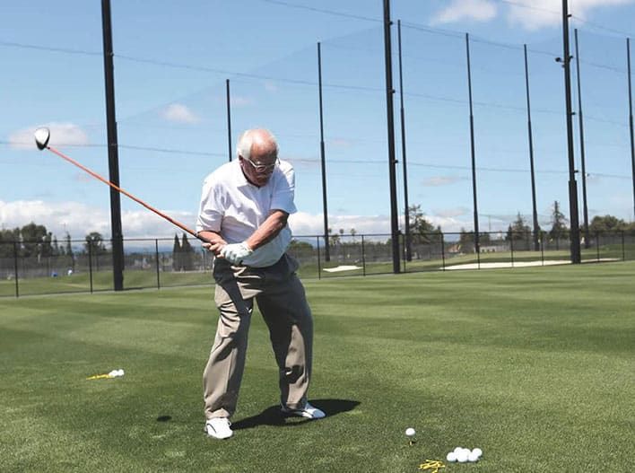 Roger Maltbie in a white shirt and beige pants swings a golf club on a grassy driving range with golf balls scattered on the ground. Tall netting and trees are visible in the background.