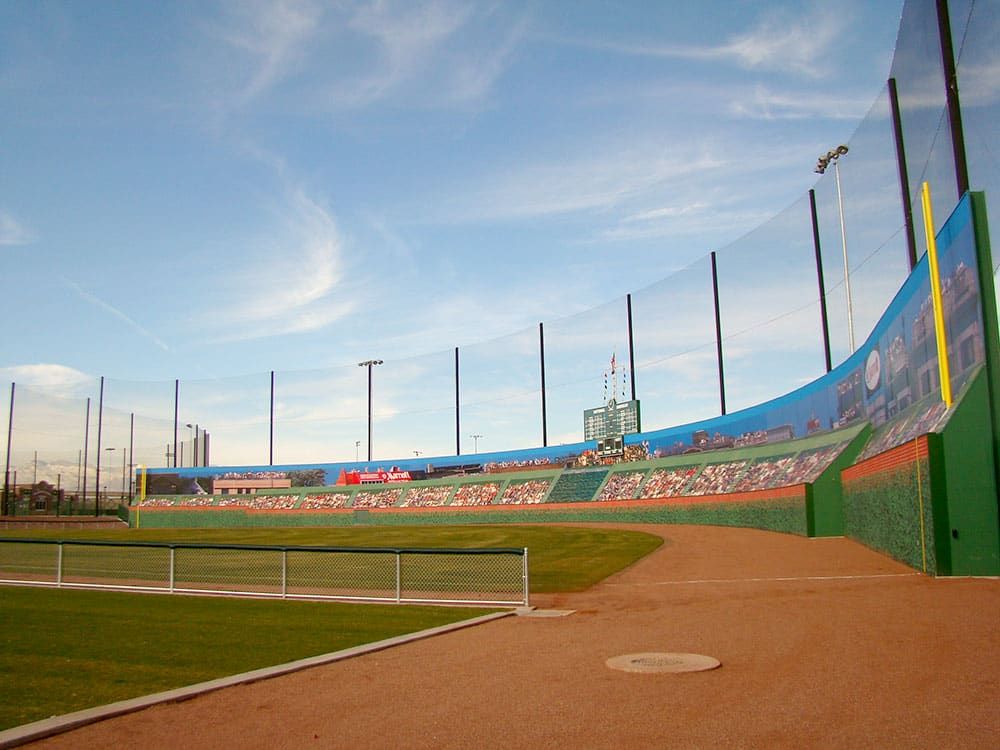 Large baseball stadium with a manicured field, a brown dirt track, and an advertisement-covered outfield wall surrounded by a tall netting system. Stadium lights and a clear sky are visible in the background.