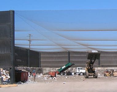 A litter transfer station with a netting roof and walls. Machinery and piles of rubbish visibile inside the station, with a dump truck and front loader tractor visible lifting a bucket.