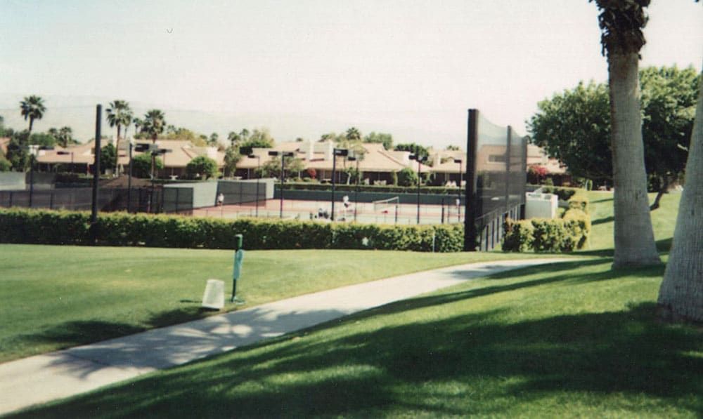 A tennis court with players is surrounded by netting fences, hedges, nearby buildings and palm trees, with a grassy foreground and pathway leading to the court.