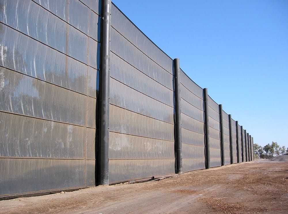 A tall, dark-colored windscreen barrier fence along a dirt road with clear blue sky in the background.