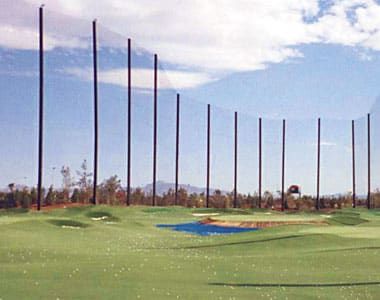 A well-maintained driving range with several tall steel poles and netting in the background. The sky is partly cloudy, and mountains are visible in the distance.