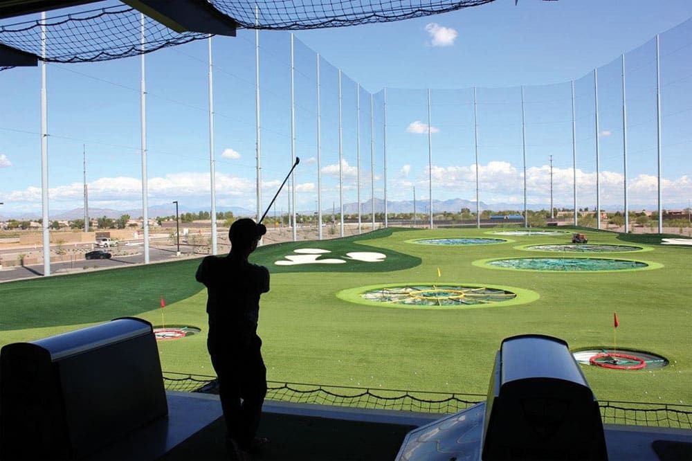 A person is swinging a golf club at Top Golf with targets on the ground. The range is surrounded by tall netting, and the sky is clear with some clouds.