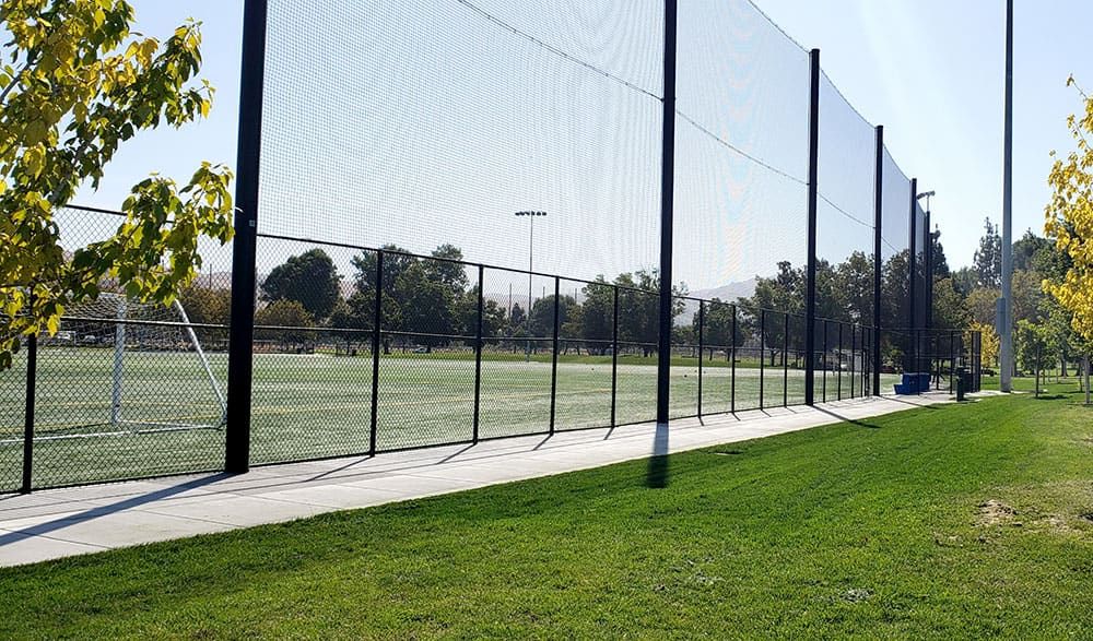 A tall netting fence surrounds a green soccer field with goal posts, surrounded by trees, under a clear sky.