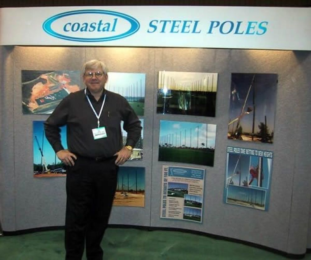 Ken Jones, Coastal Netting Systems President & CEO, stands in a PGA booth with sign reading “Coastal Steel Poles”. Photos and displays on the wall feature various steel pole images and promotional materials.