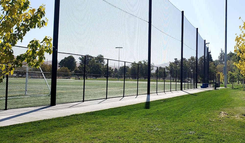 A fenced soccer field surrounded by a tall soccer netting fence and trees on a sunny day.