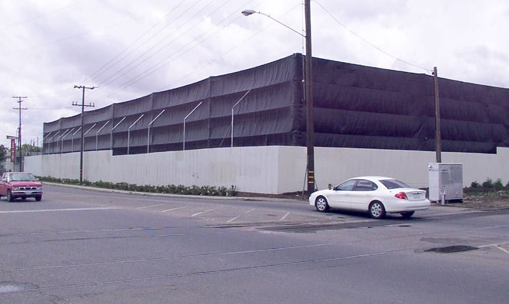 A large recycling facility covered with black netting is seen from the street. A white car is parked nearby on the right side of the image. Power lines and a lamp post are visible.