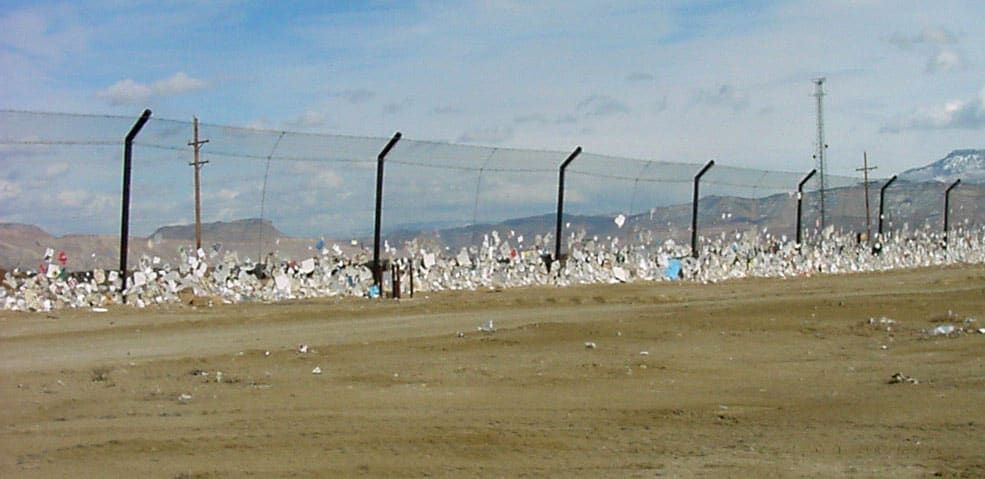 A stationary litter fence with lots of trash and debris stuck along its base stretches across a dry, barren landscape with mountains in the background.