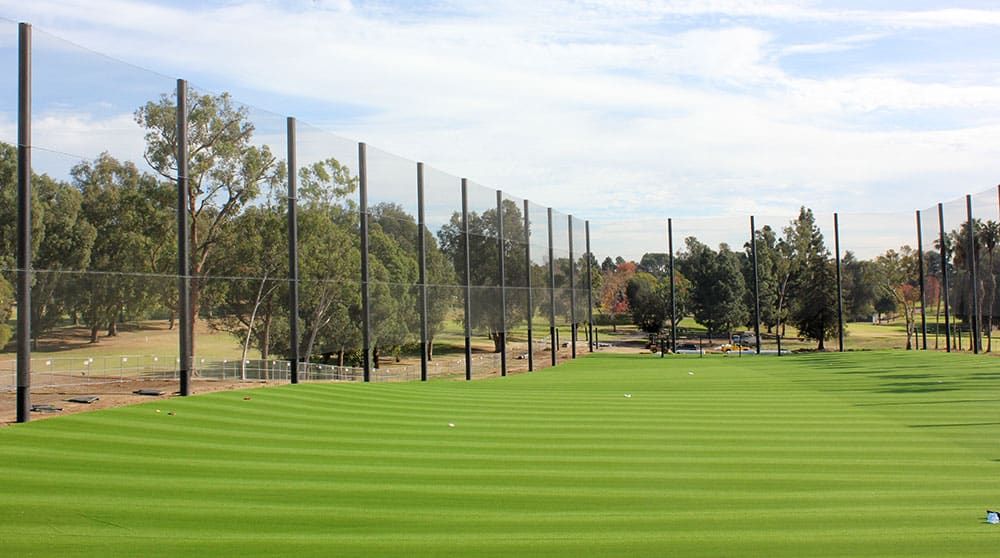 A well-maintained golf driving range with green grass and tall netting to catch balls. Trees and a bright, cloudy sky are in the background.
