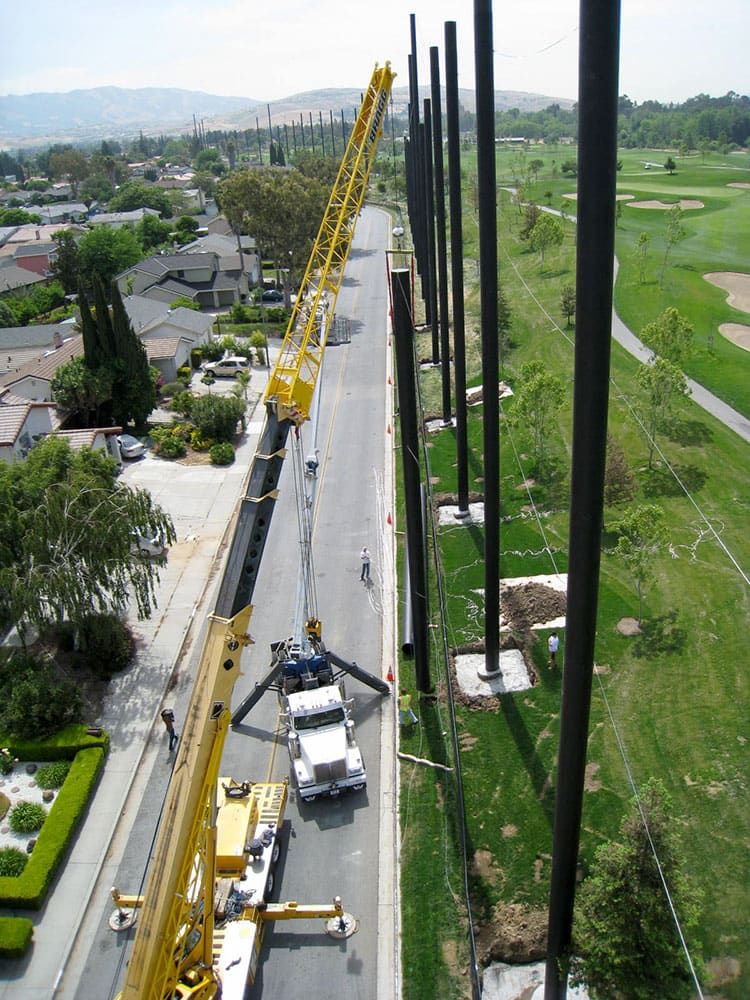 Aerial view of cranes and workers installing tall steel poles along a residential street, with a golf course on the right.