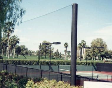 Tennis court with surrounding netting fence, palm trees, and lamp posts under a clear blue sky.