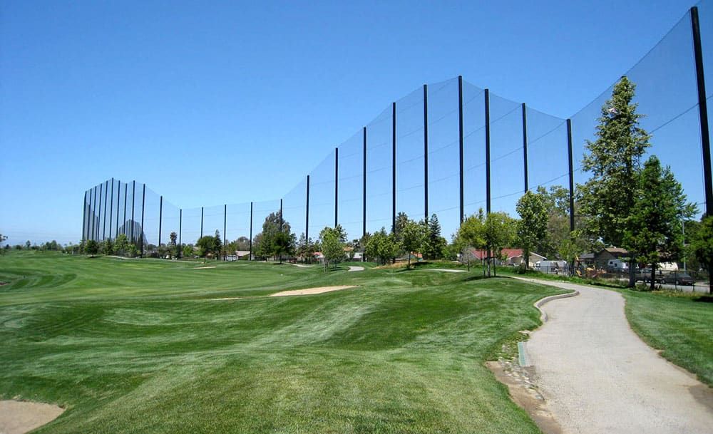 A lush green golf course with a paved golf cart path and towering protective netting along the perimeter under a clear blue sky.
