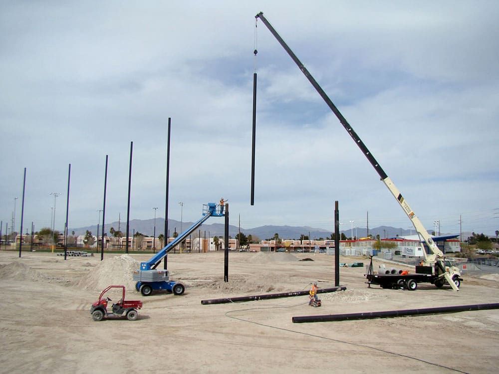 Construction site with a crane and a cherry picker lift installing a ring of tall black poles for a netting system. Several workers are present, and there is a small red vehicle nearby. Urban buildings and distant mountains are visible in the background.