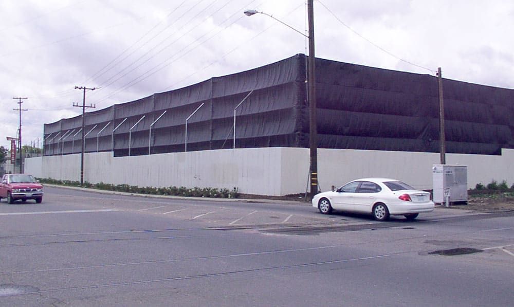 A large industrial building with black netting on top and cement walls is situated by a street corner. A white car is parked nearby under overcast skies.