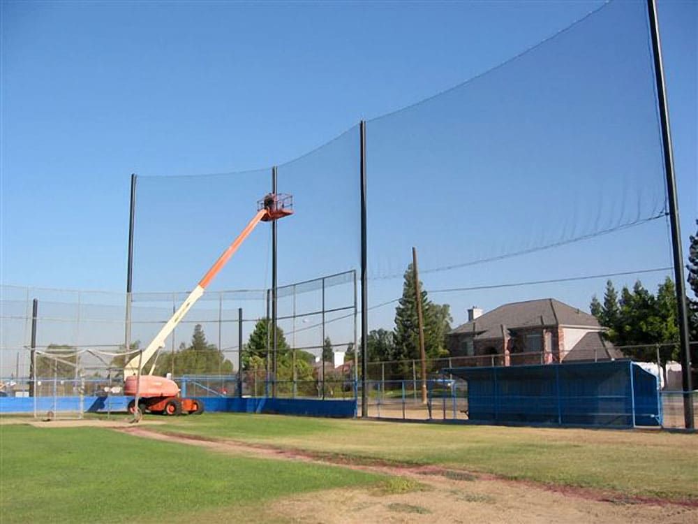 A construction lift is elevating a worker to install netting around an outdoor baseball field, with nearby houses and trees visible in the background.