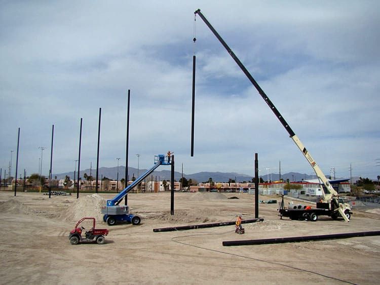 A construction site with cranes lifting Coastal’s steel poles into place, workers in hard hats, and various machinery on a dirt surface under a cloudy sky.