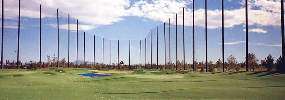 Panorama of a driving range with green turf, surrounded by tall safety netting and poles under a partly cloudy sky.