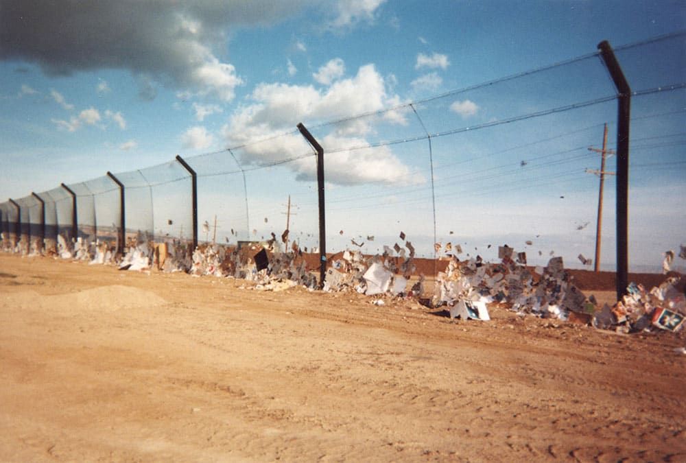 A stationary litter fence with scattered trash and debris stuck along its base, set against a dry, dirt-covered landscape and a partly cloudy sky.