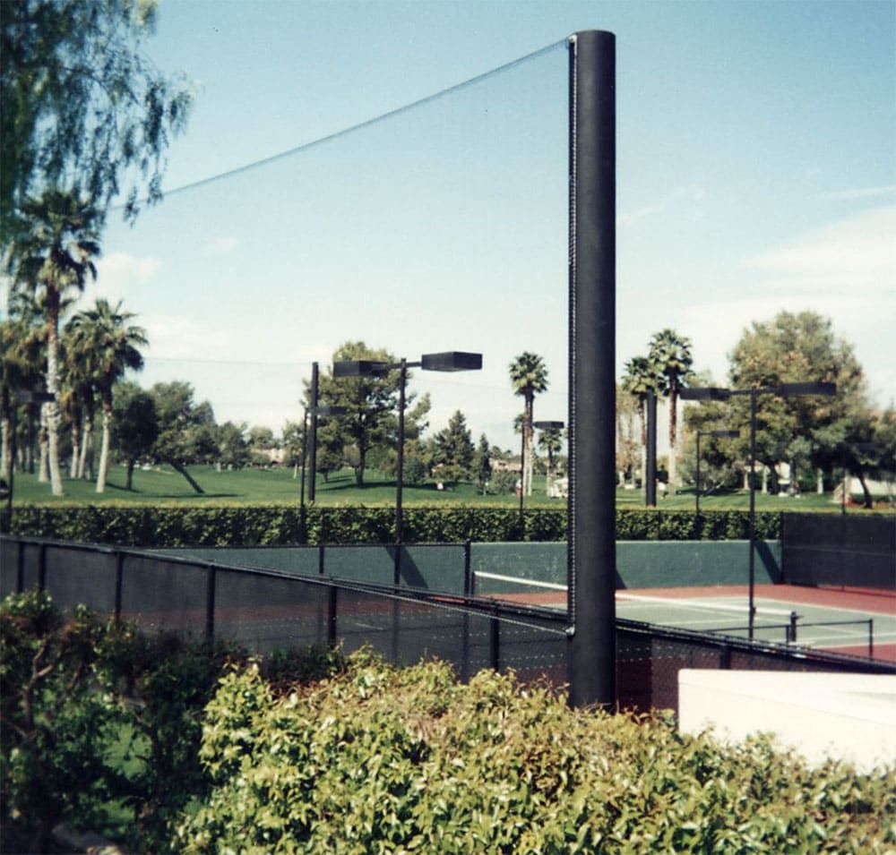 Outdoor tennis court surrounded by netting system fence, green trees and bushes, with a clear sky above and a grassy field in the background.