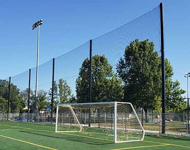 A soccer field with green grass, yellow lines, and a goal post, surrounded by a tall netting system. There are trees behind the net and a clear blue sky.