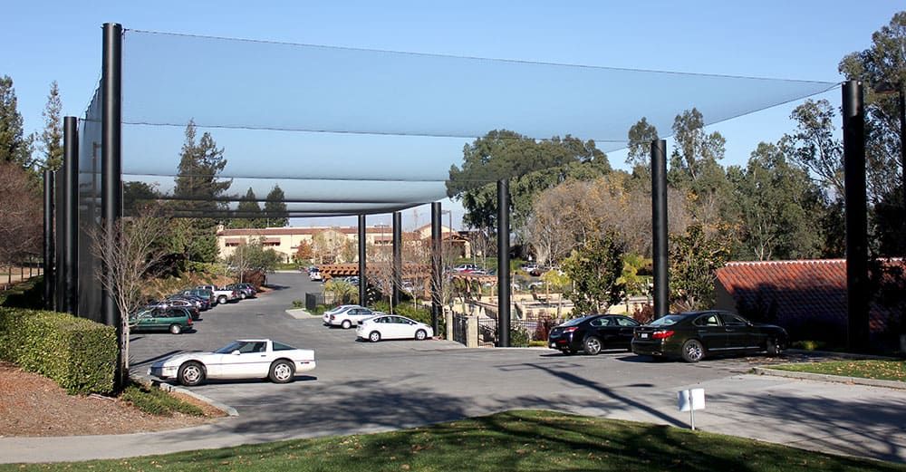 A parking lot with several cars is surrounded by tall poles supporting a large, netting structure overhead, protecting the cars from errant golf balls. Trees and buildings are visible in the background.